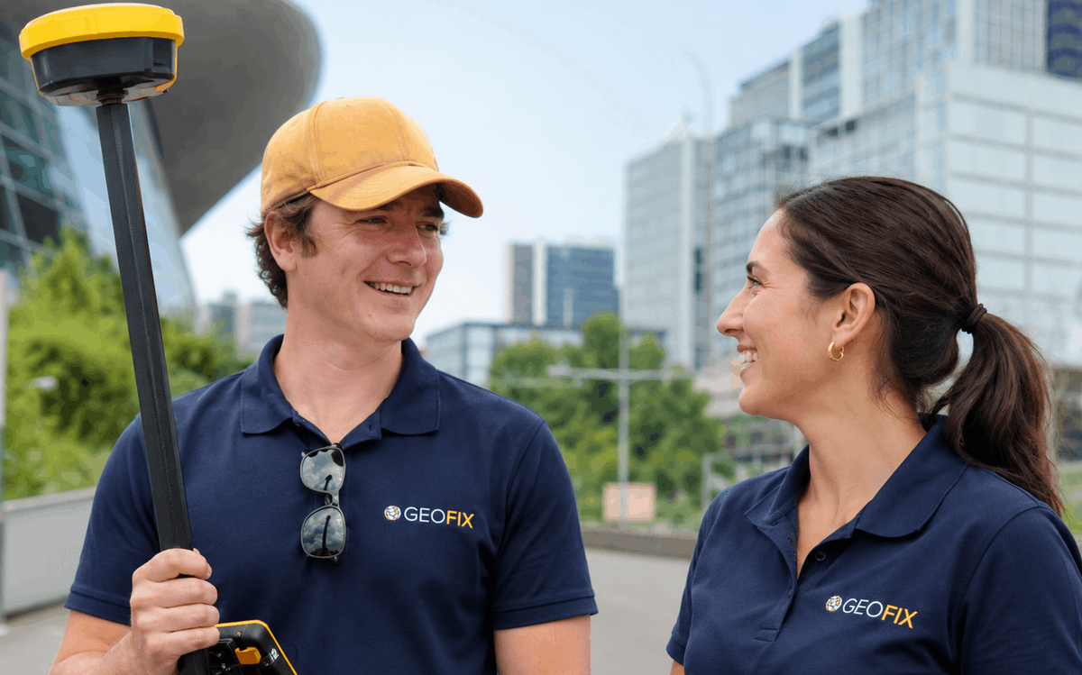 Two GEOFIX employees standing outdoors on a city street, one holding a GPS surveying pole and the other holding a tablet, both wearing navy blue polo shirts with the GEOFIX logo.